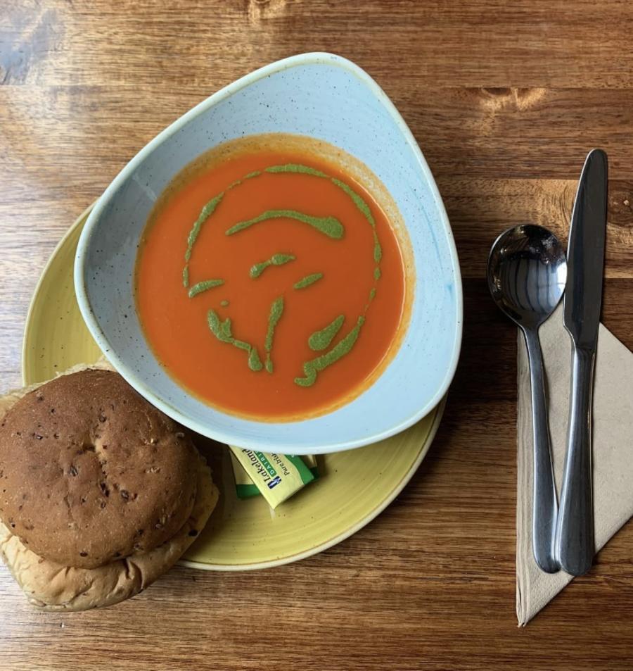 Burford on the Hill - Wonderful Winchcombe Overhead view of a place setting on a wooden table featuring a bowl of smooth tomato soup drizzled with green herb oil. The bowl sits on a light green plate, accompanied by a bread roll. To the right are a neatly placed knife and fork on a folded napkin, along with a small condiment container. The scene is warmly lit, highlighting the textures of the wood and the vibrant color of the soup.