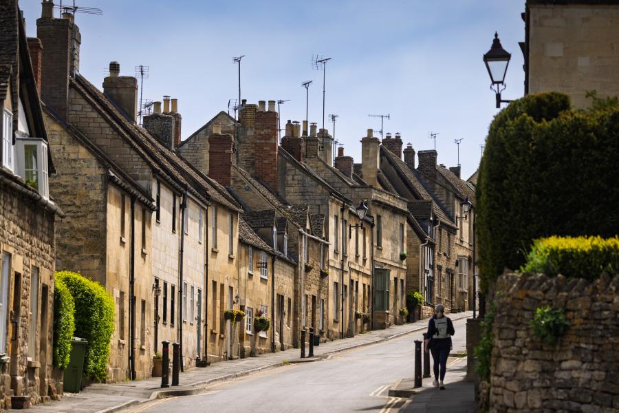 Burford on the Hill - Wonderful Winchcombe A narrow, gently curving street lined with closely packed stone houses featuring sloped roofs, chimney stacks, and small windows. The buildings vary in warm neutral tones like beige, cream, and light brown, with some greenery climbing walls and hedges along the edges. A single pedestrian walks along the pavement on the right side of the road. A low stone wall and neatly trimmed bush appear in the foreground, while traditional street lamps rise above the homes. The sky overhead is pale and clear, casting soft daylight across the quiet scene in Winchcombe.