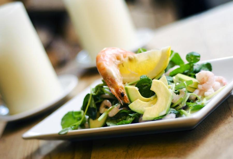 Burford on the Hill - Wonderful Winchcombe White rectangular plate holding a composed salad with sliced avocado, leafy greens, and pieces of seafood, accompanied by a lemon wedge. The dish is placed on a wooden table, with a softly blurred background featuring a white candle and dining setting, creating a warm restaurant atmosphere.