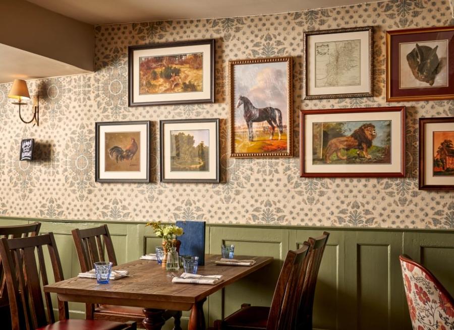 Burford on the Hill - Wonderful Winchcombe Inside the cosy restaurant at The Lion Inn. A wooden table set for four, surrounded by dark wooden chairs. The wall behind features patterned wallpaper and is decorated with a gallery of framed artwork, many depicting horses and animals in various styles and sizes. A small vase of yellow flowers sits on the table alongside menus, plates, and cutlery. Warm, ambient lighting creates an inviting, traditional atmosphere.